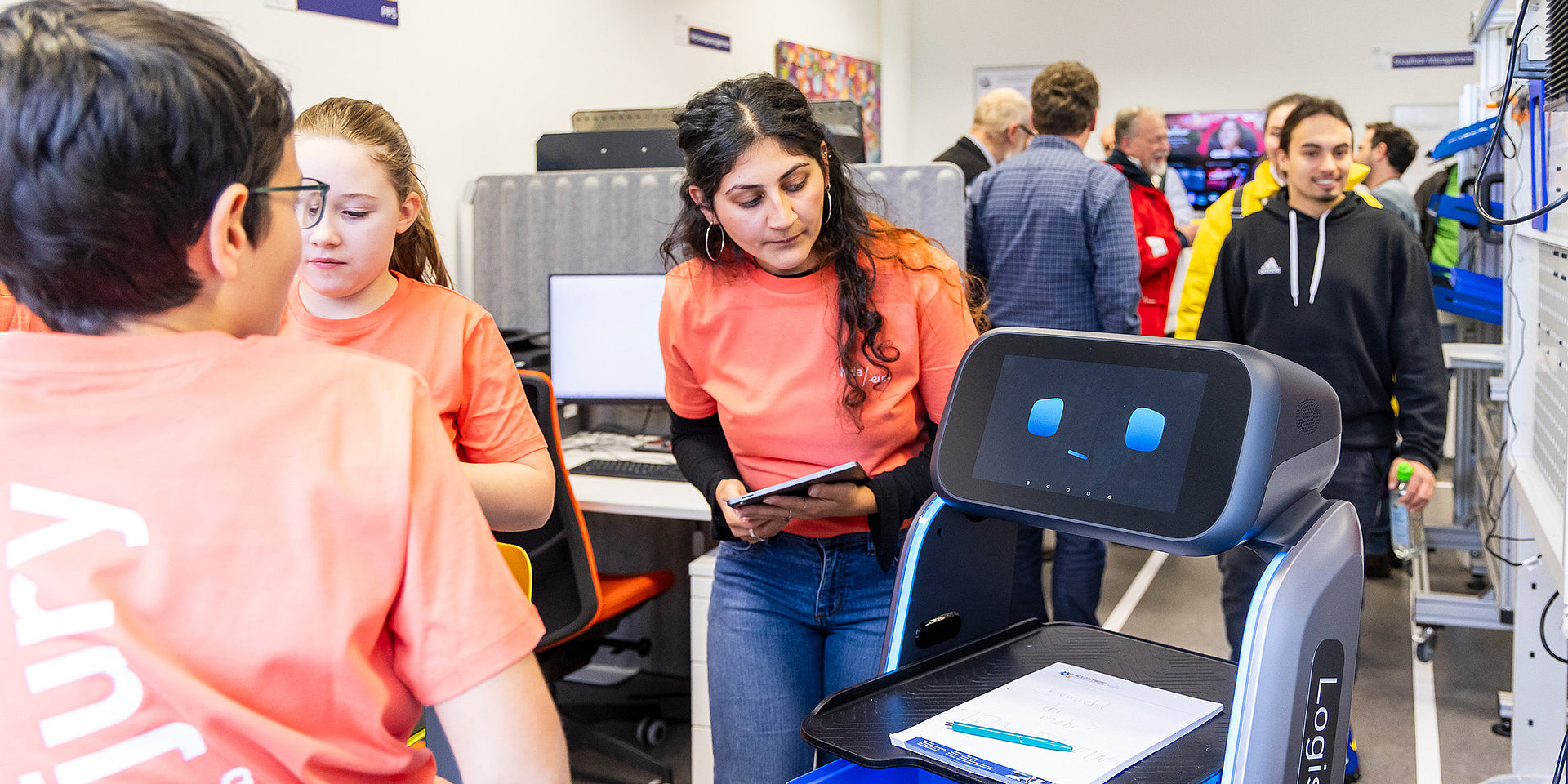 Studierende in orangenen T-Shirts im Labor mit einem kleinen Roboter.