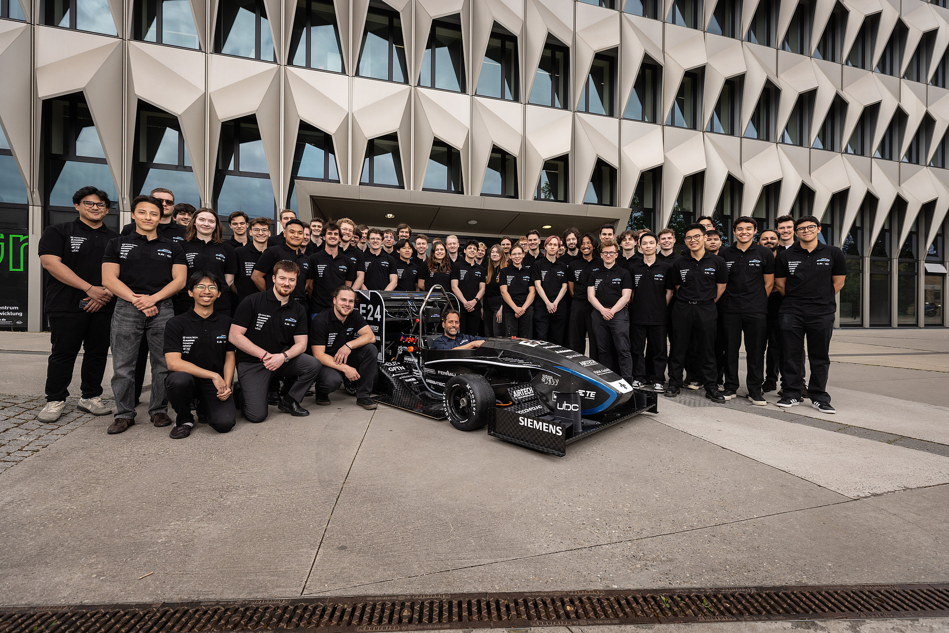 Group photo with all team members dressed in black standing behind the racing car, in front of the h_da high-rise building