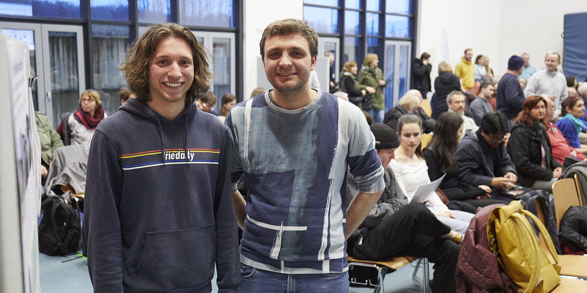 Zwei Studierende stehen nebeneinander und lächeln in die Kamera, im Hintergrund Stuhlreihen mit vielen Menschen