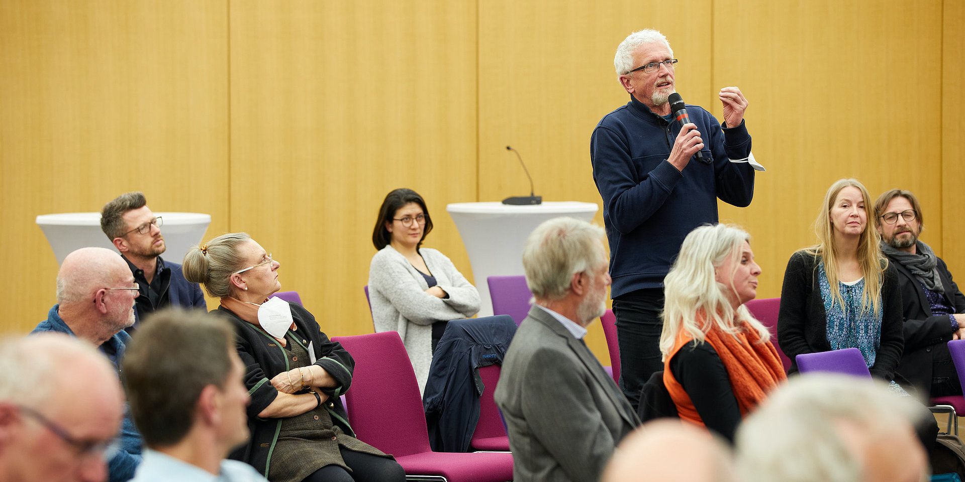 Ein Zuschauer einer Podiumsdiskussion stellt den Podiumsteilnehmern auf der Bühne Fragen.