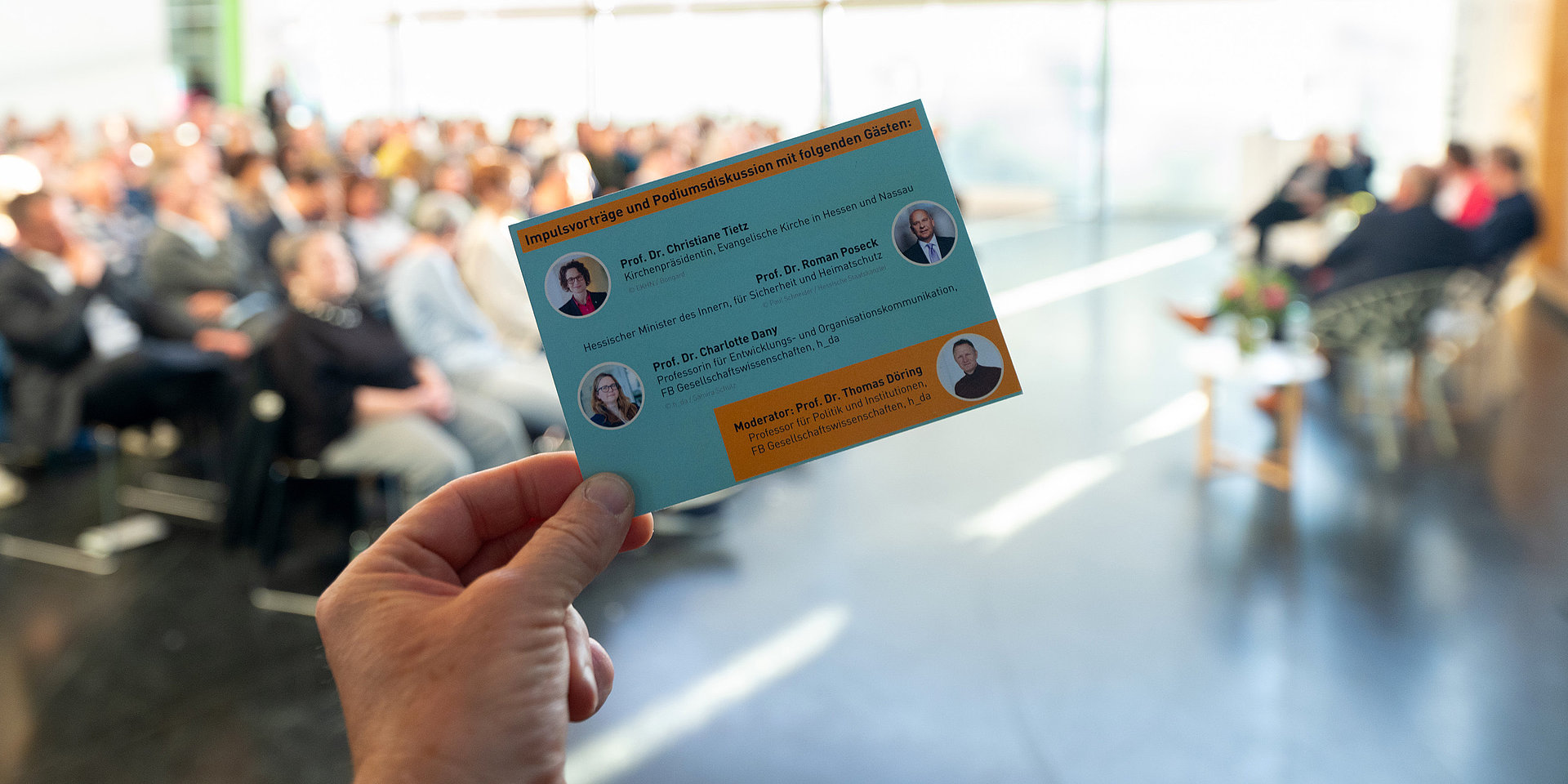 Die Einladungskarte zum Dialog-Forum im Vordergrund, dahinter unscharf das Auditorium.