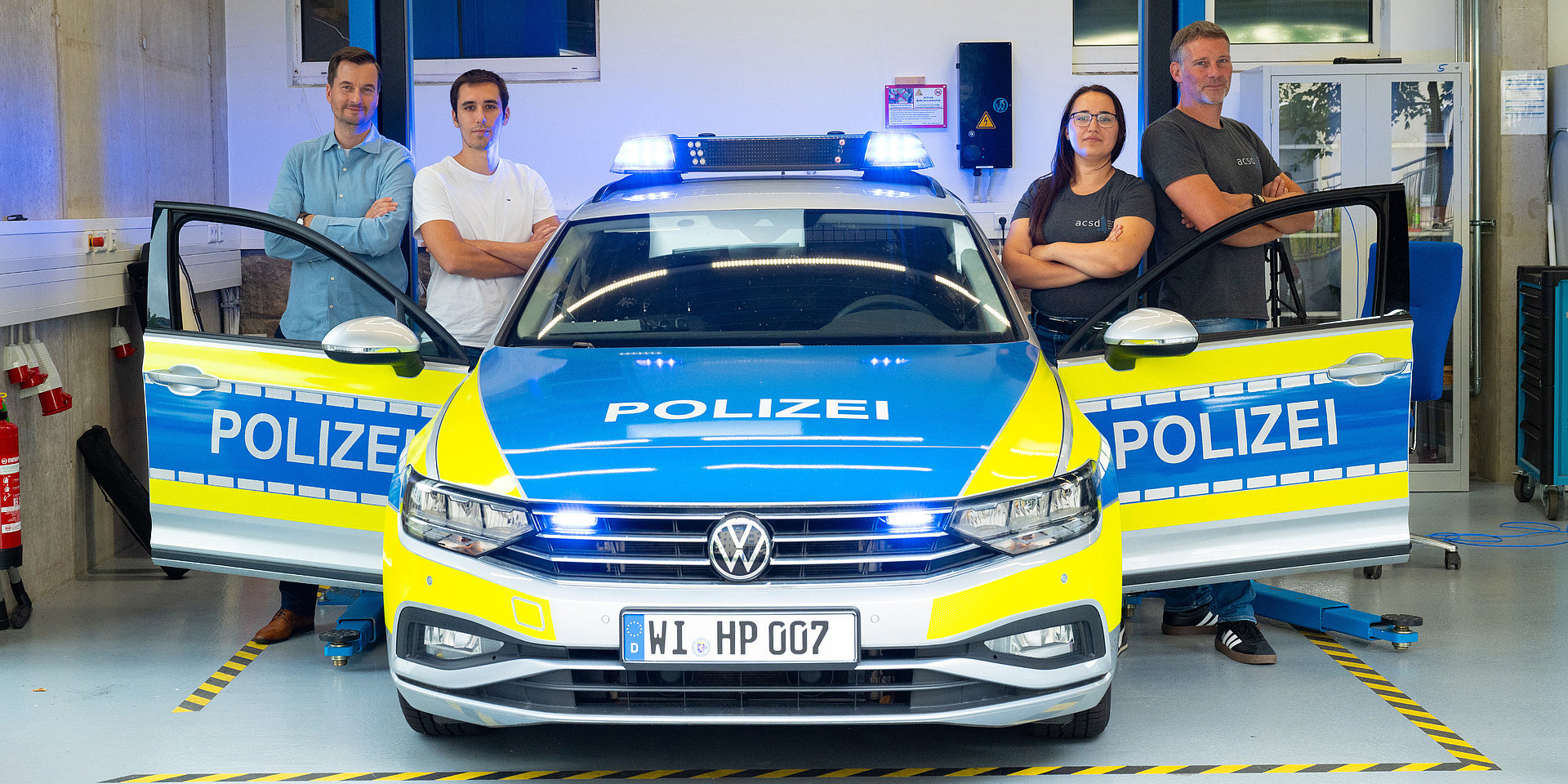 Vier Personen stehen an einem Polizeiauto und gucken in die Kamera.