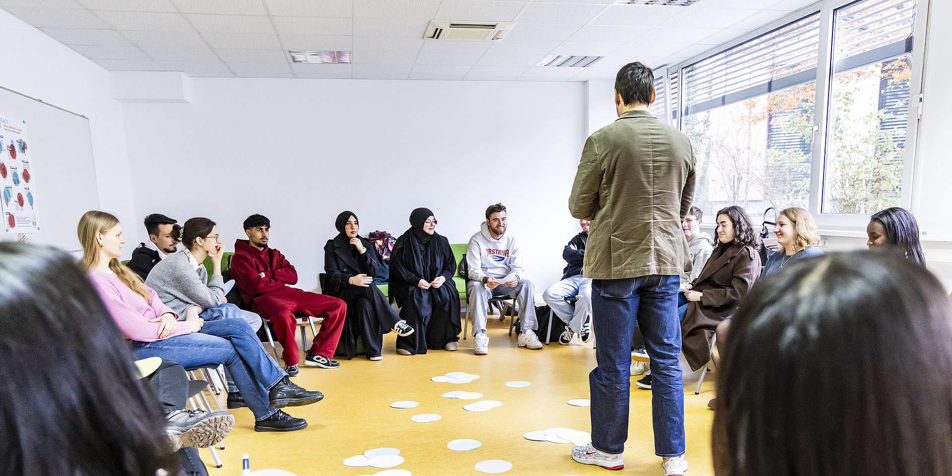 Studierende sitzen im Stuhlkreis, Trainer in der Mitte, auf dem Boden liegen beschriftete Pappschilder